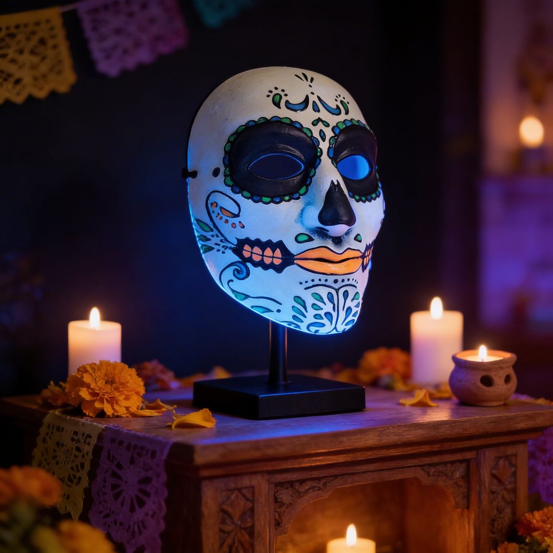 A colorful Day of the Dead mask is displayed on a wooden stand, surrounded by lit candles and flowers, creating a festive atmosphere.
